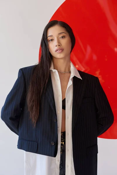 Moda adelante, bastante mujer asiática en traje de moda posando cerca de vidrio rojo sobre fondo gris, blazer y pantalones cortos de látex negro, modelo juvenil, fotografía de estudio, conceptual - foto de stock