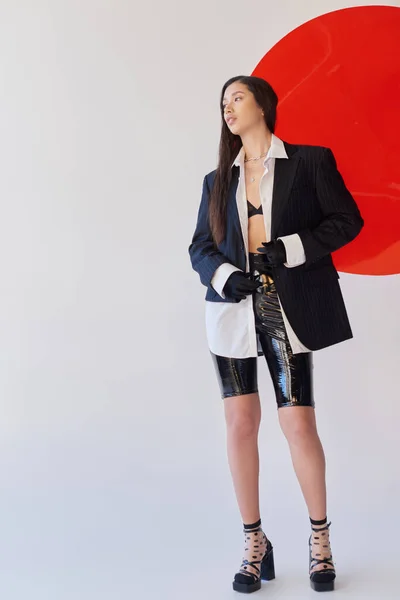 Moda para a frente, mulher asiática bonita na roupa da moda posando perto de vidro vermelho no fundo cinza, blazer e calções de látex, modelo jovem em luvas, moda e estilo, fotografia de estúdio, comprimento total — Fotografia de Stock