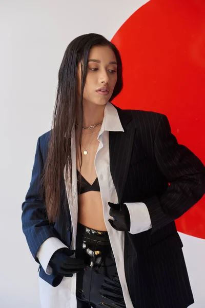 Смелый стиль, красивая азиатская женщина в модном наряде позирует возле красного стекла на сером фоне, блейзер и черные латексные шорты, молодая модель в перчатках, студийная фотография, концептуальный — стоковое фото