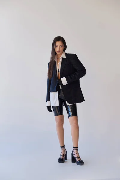 Холодный стиль, молодая женщина в трико с латексными шортами, позирующими на сером фоне, блейзером и черными перчатками, молодая модель на высоких каблуках, студийная фотография, концептуальная, полная длина — стоковое фото
