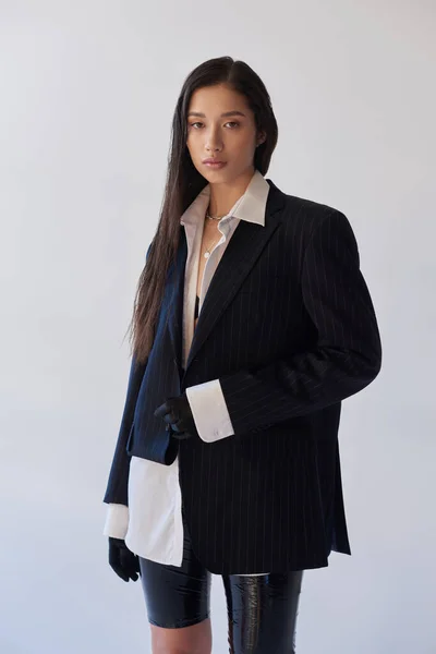 Moda juvenil, morena mujer asiática en traje de moda posando sobre fondo gris, camisa blanca, chaqueta y guantes negros, modelo joven, fotografía de estudio, conceptual - foto de stock