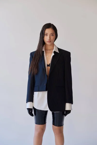 Молодая мода, брюнетка азиатская женщина в латексных шортах позирует на сером фоне, белая рубашка, пиджак и черные перчатки, молодая модель, студийная фотография, концептуальная — стоковое фото