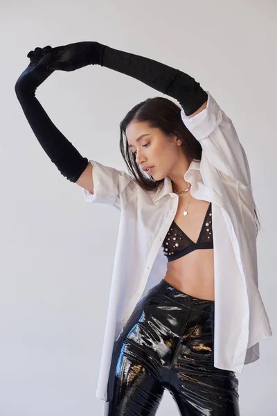 Personal style, young asian woman with brunette hair posing with raised hands on grey background, white shirt and black gloves, model posing in latex shorts in studio, fashion statement, bold style — Stock Photo
