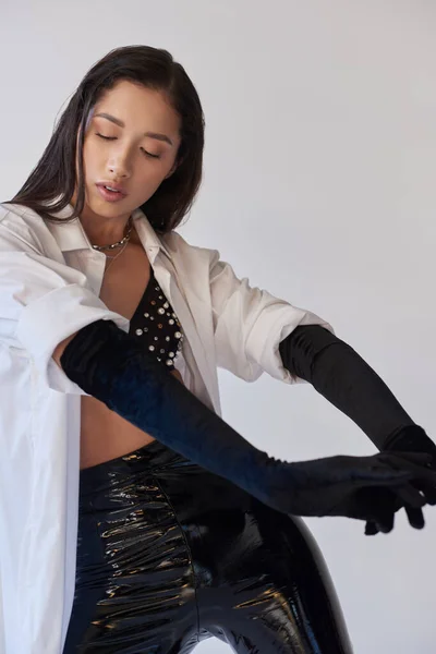 Бюстгальтер взгляд, молодая азиатка с брюнеткой волосы позируют на сером фоне, белая рубашка и черные перчатки, модель позирует в латексных шортах в студии, мода заявление, смелый стиль — стоковое фото