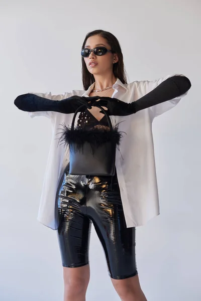 Смелый стиль, мода заявление, азиатская женщина в солнцезащитных очках позирует с пернатой сумочкой на сером фоне, молодая модель, черные перчатки и белая рубашка, латексный стиль, концептуальный, личный стиль — стоковое фото