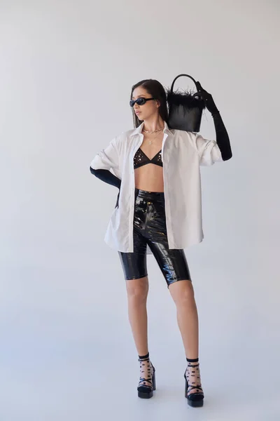 Мода заявление, азиатка в солнцезащитных очках позирует с пернатой сумочкой на сером фоне, молодая модель, черные перчатки и белая рубашка, латексные шорты, концептуальный, личный стиль — стоковое фото