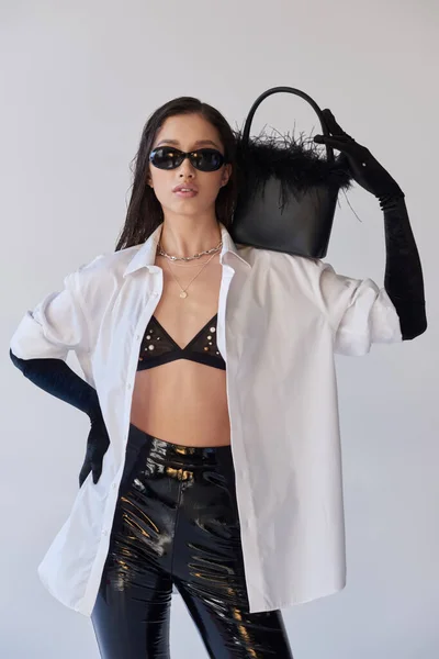 Fashion forward, asian woman in sunglasses posing with feathered handbag and hand on hip on grey background, young model, black gloves and white shirt, latex shorts, conceptual, personal style — Stock Photo