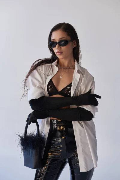 Kantigen Stil, asiatische Frau mit Sonnenbrille posiert mit gefiederter Handtasche und verschränkten Armen auf grauem Hintergrund, junges Modell, schwarze Handschuhe und weißes Hemd, Latex-Shorts, konzeptioneller, persönlicher Stil — Stockfoto