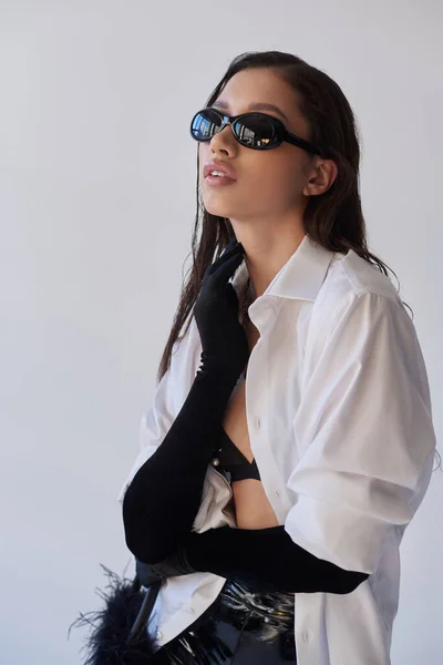 Смелый стиль, модный вид, азиатка в солнцезащитных очках позирует с пернатой сумочкой на сером фоне, молодая модель в черных перчатках и белой рубашке, концепция моды, личный стиль — стоковое фото