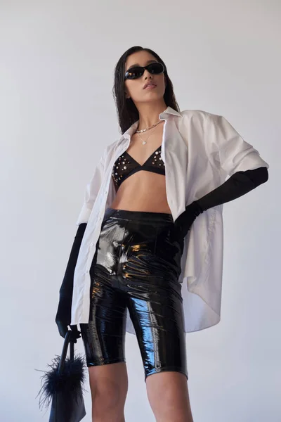 Personal style, fashion statement, asian woman in sunglasses posing with hand on hip and feathered purse on grey background, young model in latex shorts, black gloves and white shirt, conceptual — Stock Photo