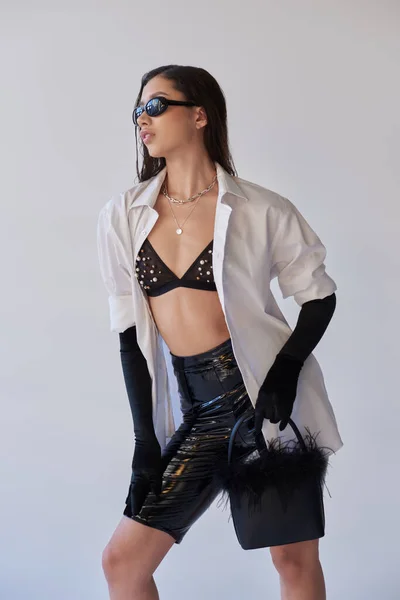 Persönlicher Stil, Mode nach vorne, asiatische Frau mit Sonnenbrille posiert mit gefiederter Handtasche auf grauem Hintergrund, junges Model in Latex-Shorts, schwarzen Handschuhen und weißem Hemd, konzeptionell — Stockfoto
