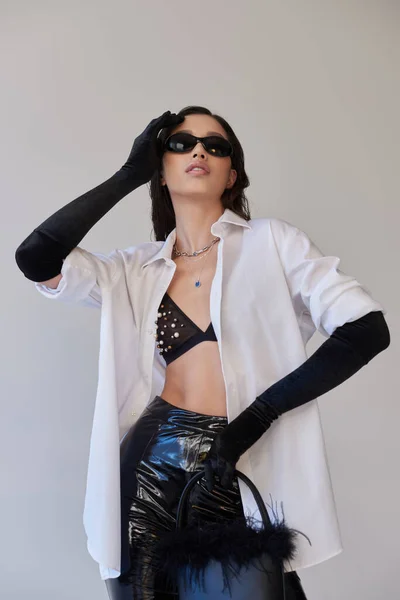 Модный вид, мода заявление, брюнетка азиатская женщина в солнцезащитных очках позирует с пернатым кошельком на сером фоне, молодая модель в латексных шортах, черные перчатки и белая рубашка, концептуальный — стоковое фото