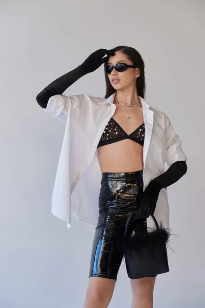 Trendy look, fashion forward, brunette asian woman in sunglasses posing with feathered purse on grey background, young model in latex shorts, black gloves and white shirt, conceptual — Stock Photo