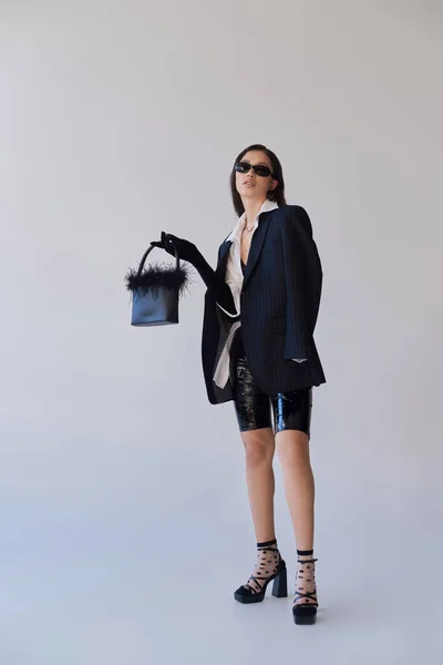 Модный вид, мода заявление, брюнетка азиатская женщина в солнцезащитных очках позирует с пернатым кошельком на сером фоне, модель в латексных шортах, черная куртка и перчатки, молодость, полная длина — стоковое фото