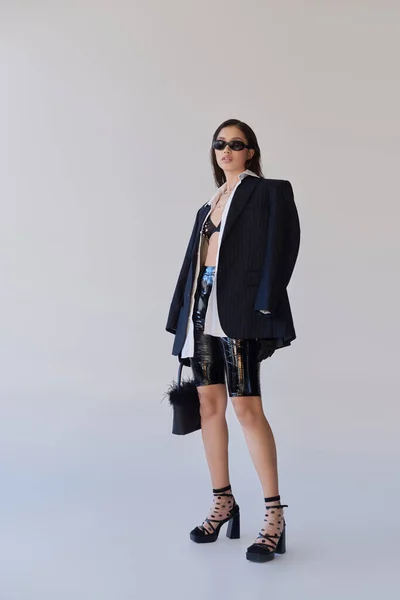 Look moderno, declaração de moda, mulher asiática morena em óculos de sol posando com bolsa de penas em fundo cinza, modelo em shorts de látex, sutiã, jaqueta preta e luvas, juventude, comprimento total — Fotografia de Stock