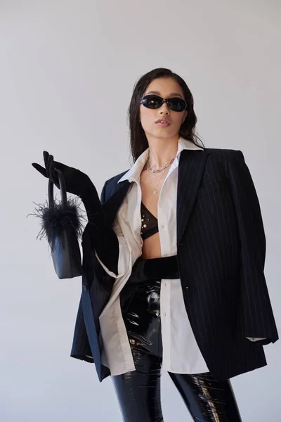 Модний наряд, особистий стиль, брюнетка азіатська жінка в сонцезахисних окулярах позує з пернатим гаманцем на сірому фоні, модель в латексних шортах, бюстгальтер, чорний піджак і рукавички, молодь і стиль — стокове фото