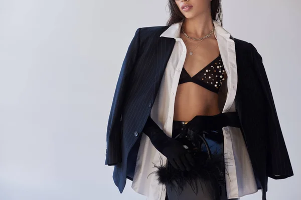 Personal style, brunette woman in bra posing with feathered purse on grey background, young model in latex shorts, blazer and black gloves, youth and style, cropped image — Stock Photo