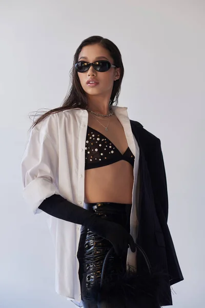 Personal style, brunette asian woman in dark sunglasses posing with blazer on grey background, young model in latex shorts, bra, white shirt and black gloves, style and trends — Stock Photo
