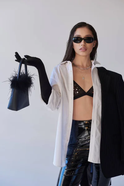 Estilo vanguardista, fotografía de estudio, mujer asiática joven en aspecto elegante y gafas de sol posando con bolso de plumas sobre fondo gris, modelo en blazer y pantalones cortos de látex, moda juvenil - foto de stock