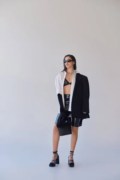 Cooler Stil, Studiofotografie, junge asiatische Frau in stylischem Look und Sonnenbrille posiert mit gefiederter Handtasche auf grauem Hintergrund, Blazer und Latex-Shorts, jugendliche Mode, volle Länge — Stockfoto