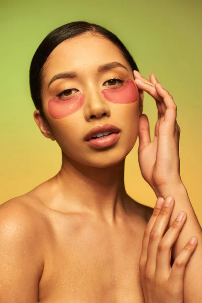 Bella e bruna donna asiatica in posa con macchie sugli occhi su sfondo verde, pendenza, bellezza, pelle disintossicante, trattamento viso, spalle nude, routine di cura della pelle, pelle luminosa, ritratto — Foto stock