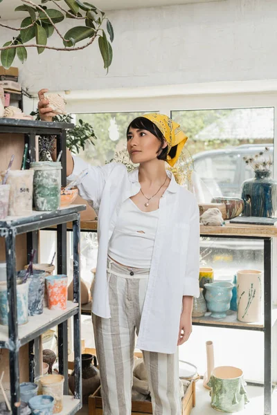 Jeune artiste asiatique en vêtements de travail et foulard prenant des produits en argile de l'étagère tout en se tenant dans la classe de poterie floue à l'arrière-plan, atelier de poterie avec artisan au travail — Photo de stock