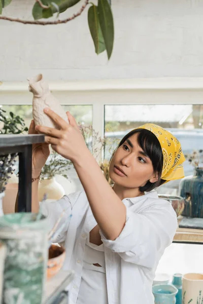 Young asian female artist in headscarf and workwear taking clay product from shelf while standing and working in blurred pottery class, pottery studio with artisan at work — Stock Photo