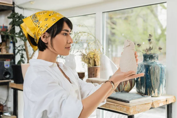 Молодая азиатская художница в рабочей одежде и платке держит глиняную скульптуру, стоя и работая в размытой керамической мастерской, гончарной мастерской с ремесленником за работой — стоковое фото