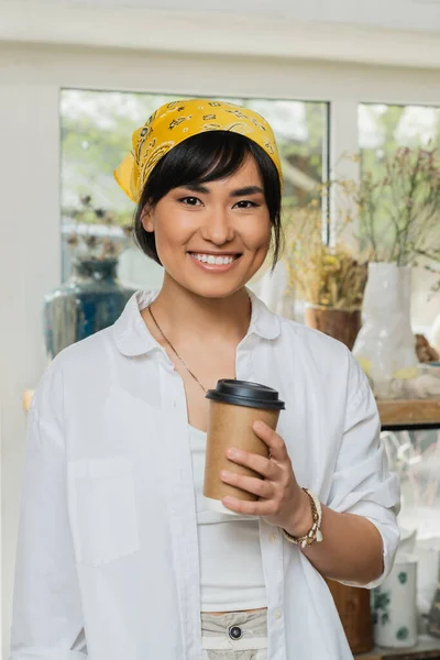 Портрет усміхненої молодої азіатки-художниці в хустці і робочому одязі, дивлячись на камеру і тримаючи каву, щоб піти, стоячи в розмитому класі кераміки, керамічна майстерня з кваліфікованим майстром — стокове фото