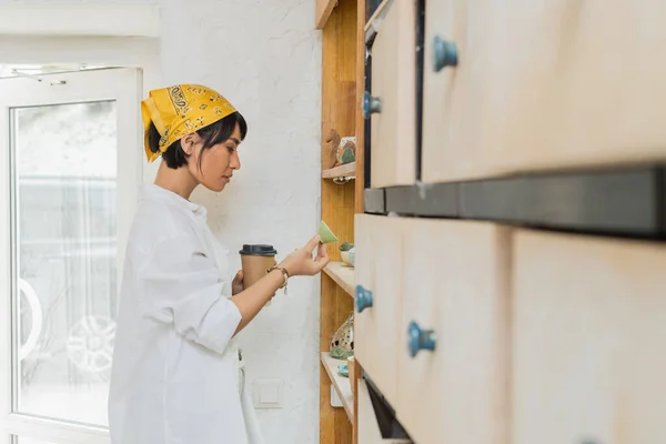 Side view of young asian female artist in headscarf and workwear holding takeaway coffee and clay product near shelves in pottery workshop, pottery workshop with skilled artisan — Stock Photo