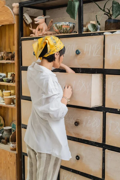 Young asian female ceramicist in headscarf and workwear opening cupboard while working and standing near clay products on shelves in ceramic studio, pottery workshop with skilled artisan — Stock Photo