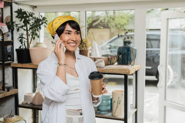 Lächelnde junge asiatische Künstlerin in Kopftuch und Arbeitskleidung, die mit dem Smartphone spricht, Kaffee to go hält und in einer verschwommenen Keramikwerkstatt steht, Kunsthandwerkerin im Töpferatelier, die sich auf die Schöpfung konzentriert — Stockfoto