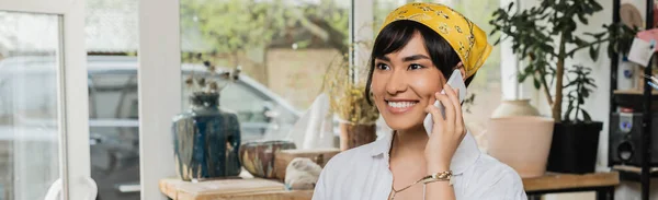 Smiling young asian craftswoman in workwear and headscarf talking on smartphone and standing n blurred ceramic workshop, artisan in pottery studio focusing on creation, banner — Stock Photo