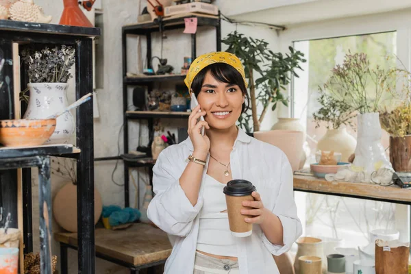 Весела молода азіатська жінка-гончарка в головних уборах і робочому одязі розмовляє на смартфоні і тримає каву, щоб піти і стояти в розмитій керамічній майстерні, майстер в керамічній студії, фокусуючись на створенні — стокове фото