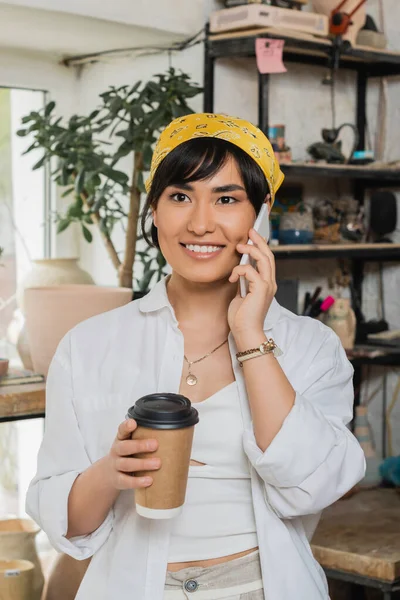 Retrato de alegre joven alfarera asiática sosteniendo café para ir y hablar en el teléfono inteligente mientras trabaja en estudio de cerámica borrosa, artesano en estudio de cerámica se centra en la creación - foto de stock