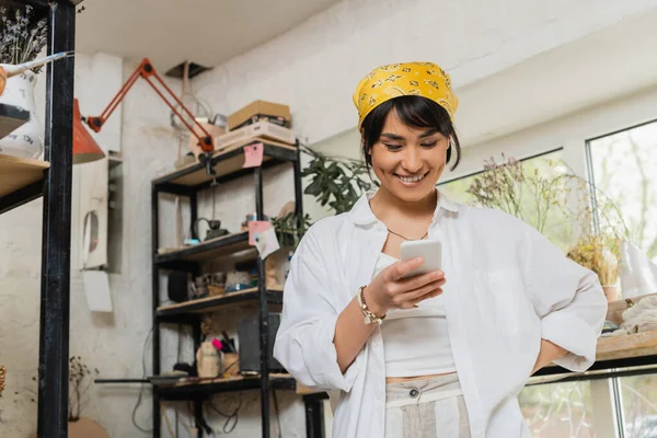Positive young asian female potter in headscarf and workwear using smartphone while standing and working in blurred ceramic workshop, artisan in pottery studio focusing on creation — Stock Photo