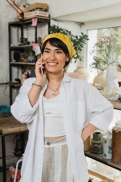 Усміхнений молодий азіатський жіночий гончар у хустці та робочому одязі розмовляє на смартфоні та тримає руку на стегні, стоячи в розмитій керамічній майстерні, майстер в керамічній студії, зосереджуючись на створенні — стокове фото
