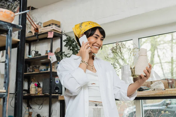 Cheerful young asian female craftswoman in headscarf and workwear talking on smartphone and holding clay sculpture while standing in ceramic workshop, artisan in pottery studio focusing on creation — Stock Photo