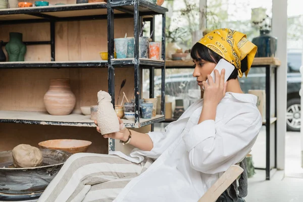 Side view of young asian artist in headscarf and workwear talking on smartphone and holding ceramic sculpture while sitting in ceramic workshop, artisan in pottery studio focusing on creation — Stock Photo