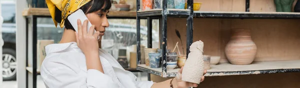 Side view of brunette asian potter in headscarf and workwear talking on smartphone and holding clay sculpture while working in ceramic studio, artisan in pottery studio focusing on creation, banner — Stock Photo