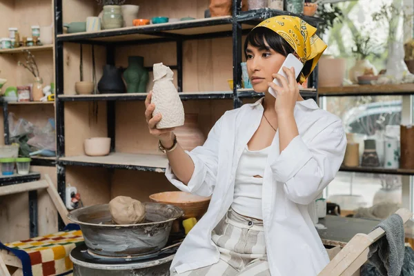 Junge asiatische Künstlerin in Kopftuch und Arbeitskleidung in Kopftuch und Arbeitskleidung spricht über Smartphone und hält Tonskulpturen in verschwommener Keramikwerkstatt, Kunsthandwerker im Töpferatelier konzentriert sich auf die Schöpfung — Stockfoto