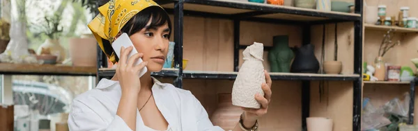 Young asian female potter in headscarf and workwear talking on smartphone and holding clay sculpture while working in ceramic workshop, artisan in pottery studio focusing on creation, banner — Stock Photo