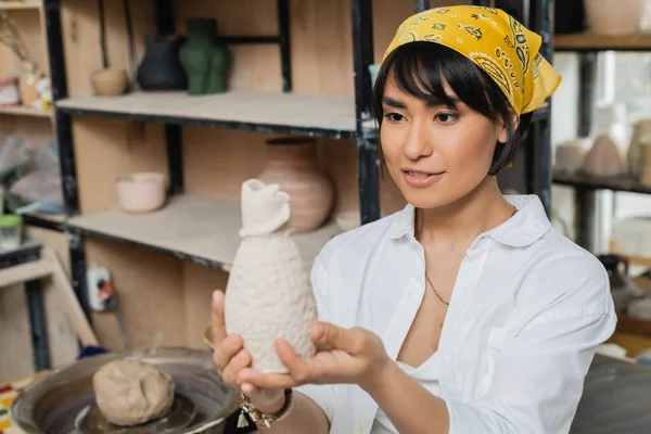 Smiling young asian artist in headscarf and workwear holding and looking at clay sculpture while working in ceramic workshop, artisan in pottery studio focusing on creation — Stock Photo