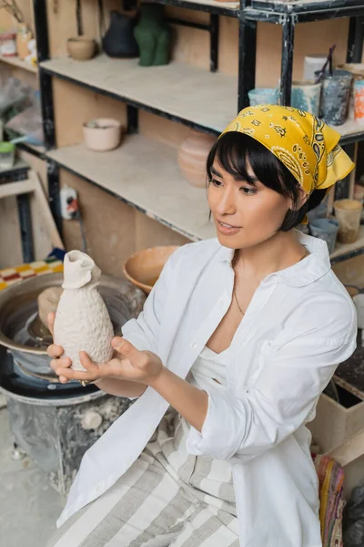 Junge asiatische Töpferin mit Kopftuch und Arbeitskleidung, die Tonerzeugnisse hält, während sie im Hintergrund in der Keramikwerkstatt sitzt und arbeitet, handwerkliches Geschick in der Töpferei — Stockfoto