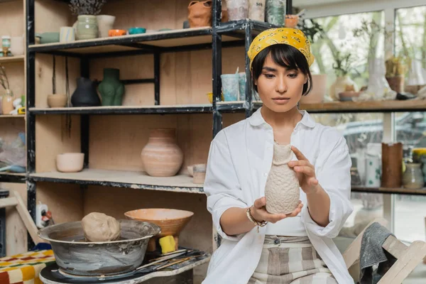 Young asian brunette female potter in headscarf and workwear holding clay sculpture near pottery wheel while working in ceramic workshop, craftsmanship in pottery making — Stock Photo