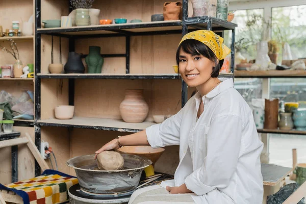 Cheerful brunette asian female potter in headscarf and workwear holding clay and looking at camera near pottery wheel in ceramic workshop, craftsmanship in pottery making — Stock Photo
