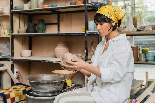 Молодая азиатская женщина-ремесленница в головном уборе и рабочей одежде держит глину во время работы рядом с гончарным колесом в керамической мастерской на заднем плане, мастерство в гончарном деле — стоковое фото