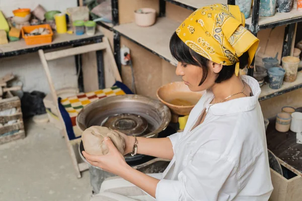 Hochwinkelaufnahme der jungen brünetten asiatischen Kunsthandwerkerin mit Kopftuch, die Ton hält und in der Nähe der Töpferscheibe und im Hintergrund im Atelier arbeitet, Handwerkskunst in der Töpferei — Stockfoto