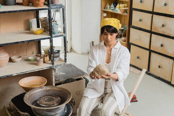 Young brunette asian female potter in workwear and headscarf holding clay near pottery wheel while working in blurred ceramic workshop, craftsmanship in pottery making — Stock Photo