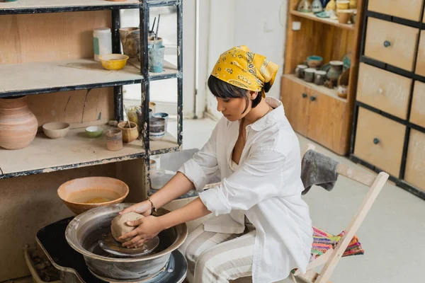 Junge asiatische Kunsthandwerkerin mit Kopftuch und Arbeitskleidung, die Ton auf Töpferscheibe formt und in der Nähe von Töpferwerkzeugen in einer verschwommenen Keramikwerkstatt arbeitet, Kunsthandwerk in der Töpferei — Stockfoto
